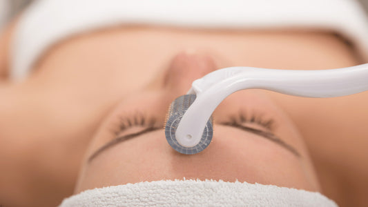 Närbild av ett ansikte under en hudbehandling där en dermaroller förs över pannan; personen ligger ner med slutna ögon och har en vit handduk runt huvudet.